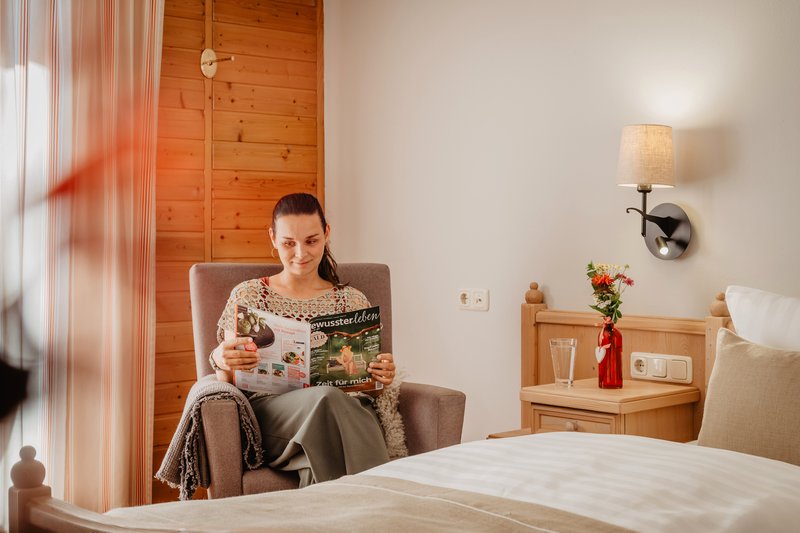 Junge Frau sitzt gemütlich im Sessel im Doppelzimmer des Hotels Sankt Leonhard und liest ein Magazin, umgeben von hellen Naturholzmöbeln und Holzwand.